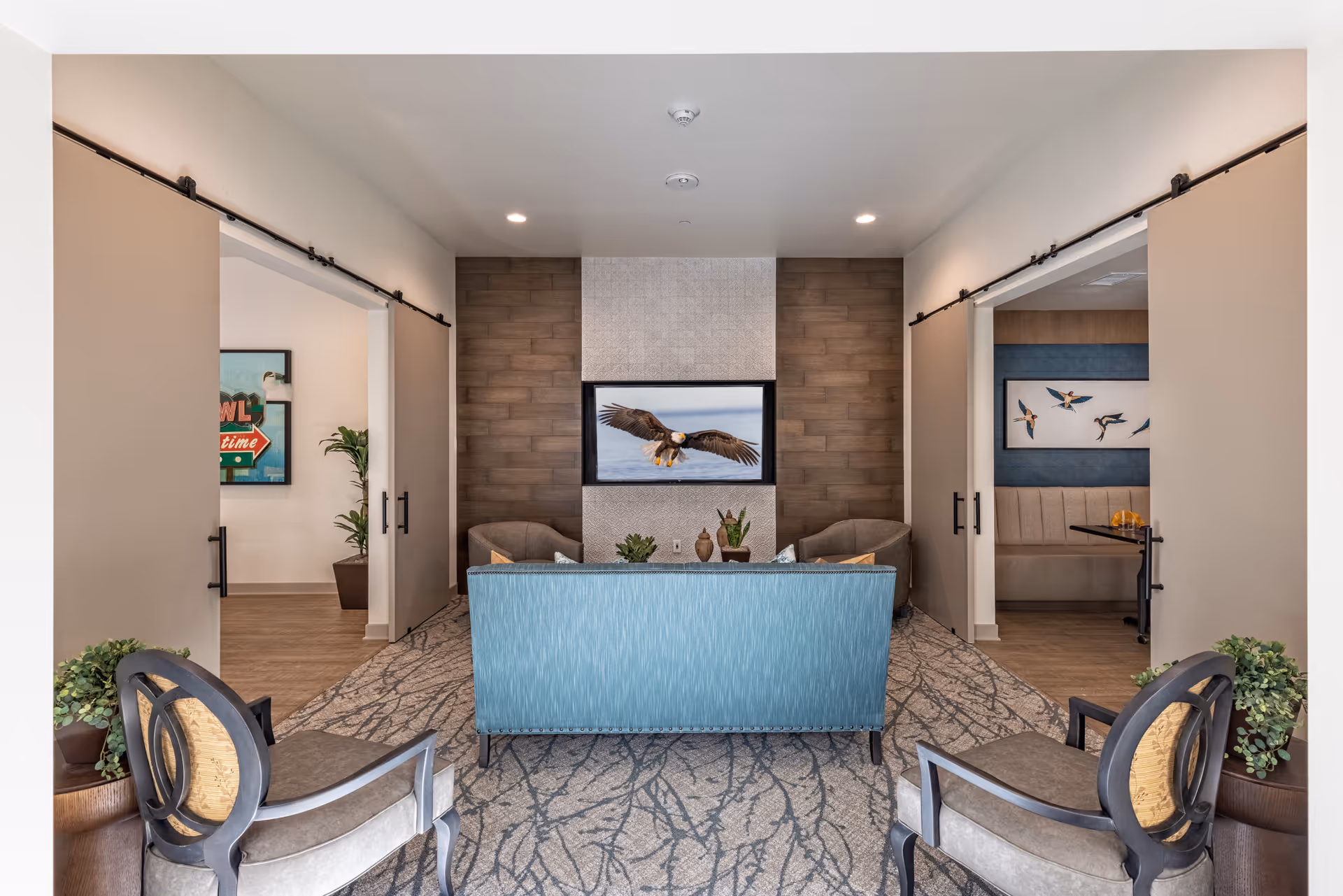 A cozy living room area with a blue sofa facing a wall-mounted TV displaying an eagle in flight. The room has two armchairs on either side of the sofa and two sliding barn doors on each side leading to other rooms. The walls are decorated with wood paneling and artwork, and there are potted plants placed around the seating area.