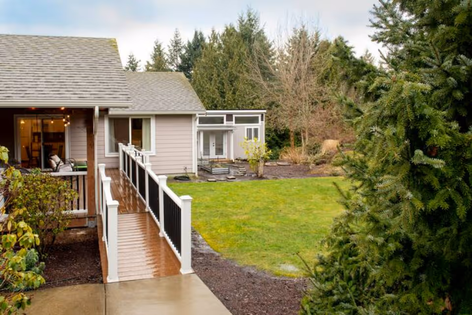 View of an outdoor area at Agape Care Adult Family Home showing a wet wooden ramp with white railings leading to a covered porch attached to a beige building. The scene includes green grass, trees, and shrubs under a cloudy sky.