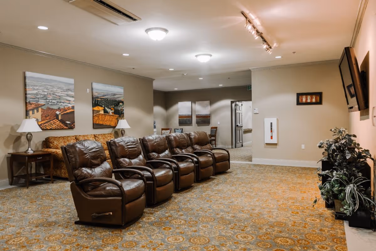 A cozy lounge area with five brown leather recliners arranged in a row facing a wall-mounted TV. Behind the recliners is a patterned sofa with two table lamps on either side. The walls are decorated with landscape paintings, and there are plants on the right side near the TV. The room has warm lighting and a patterned carpet.