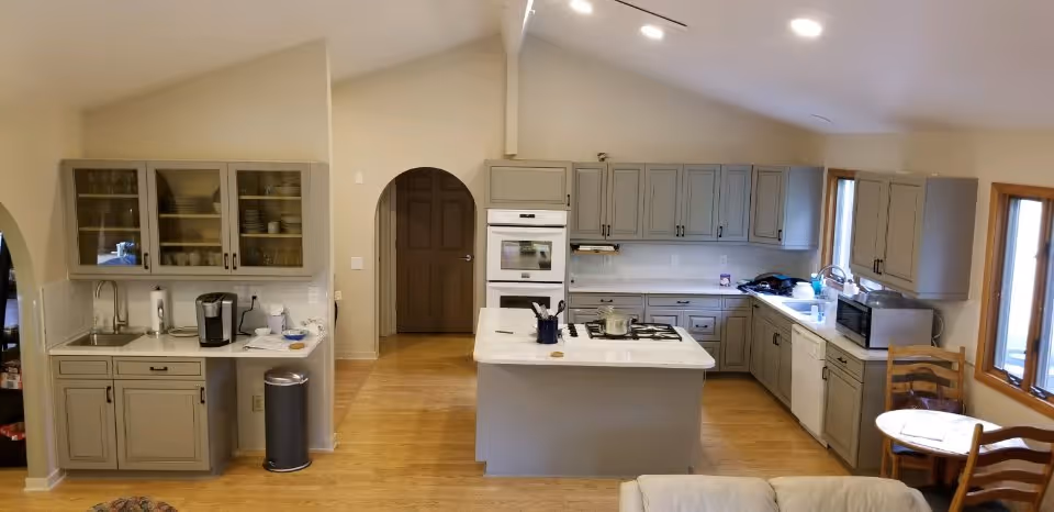 A spacious kitchen with light gray cabinets, a central island with a stove, a double oven built into the cabinetry, a sink under a window, a microwave, and a small round table with two wooden chairs near the windows. The floor is wooden, and the walls are painted a light beige color.
