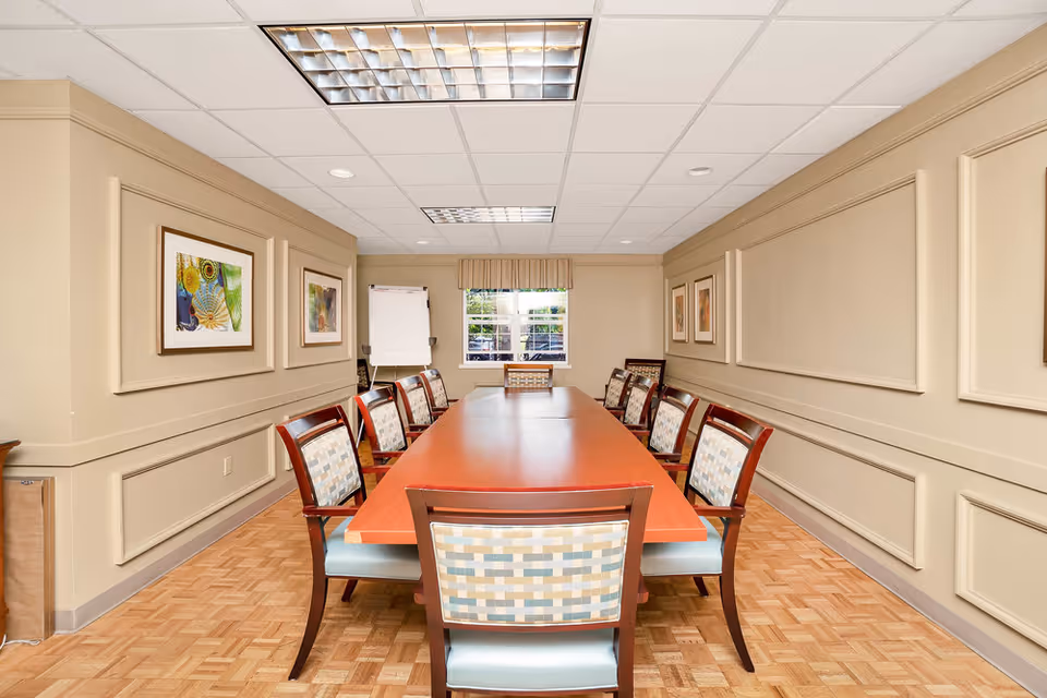 Long conference-style table with multiple chairs in a beige meeting room with framed artwork and a window at the far end.