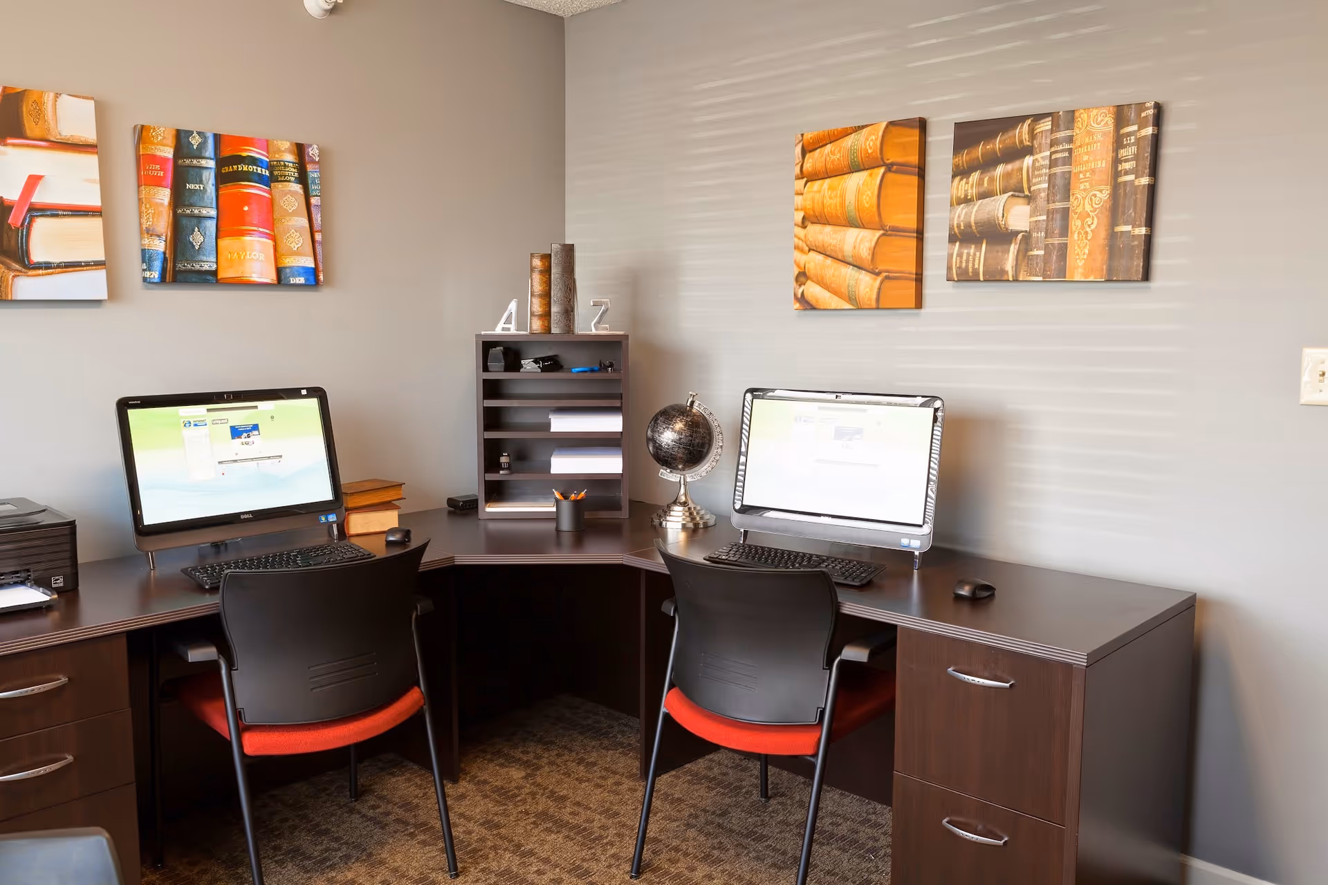 A small office area with two computer workstations on a dark wood L-shaped desk. Each workstation has a black chair with a red cushion, a keyboard, and a mouse. On the desk are a printer, a small globe, a pencil holder, and some decorative items. The walls are painted gray and decorated with framed pictures of old books.