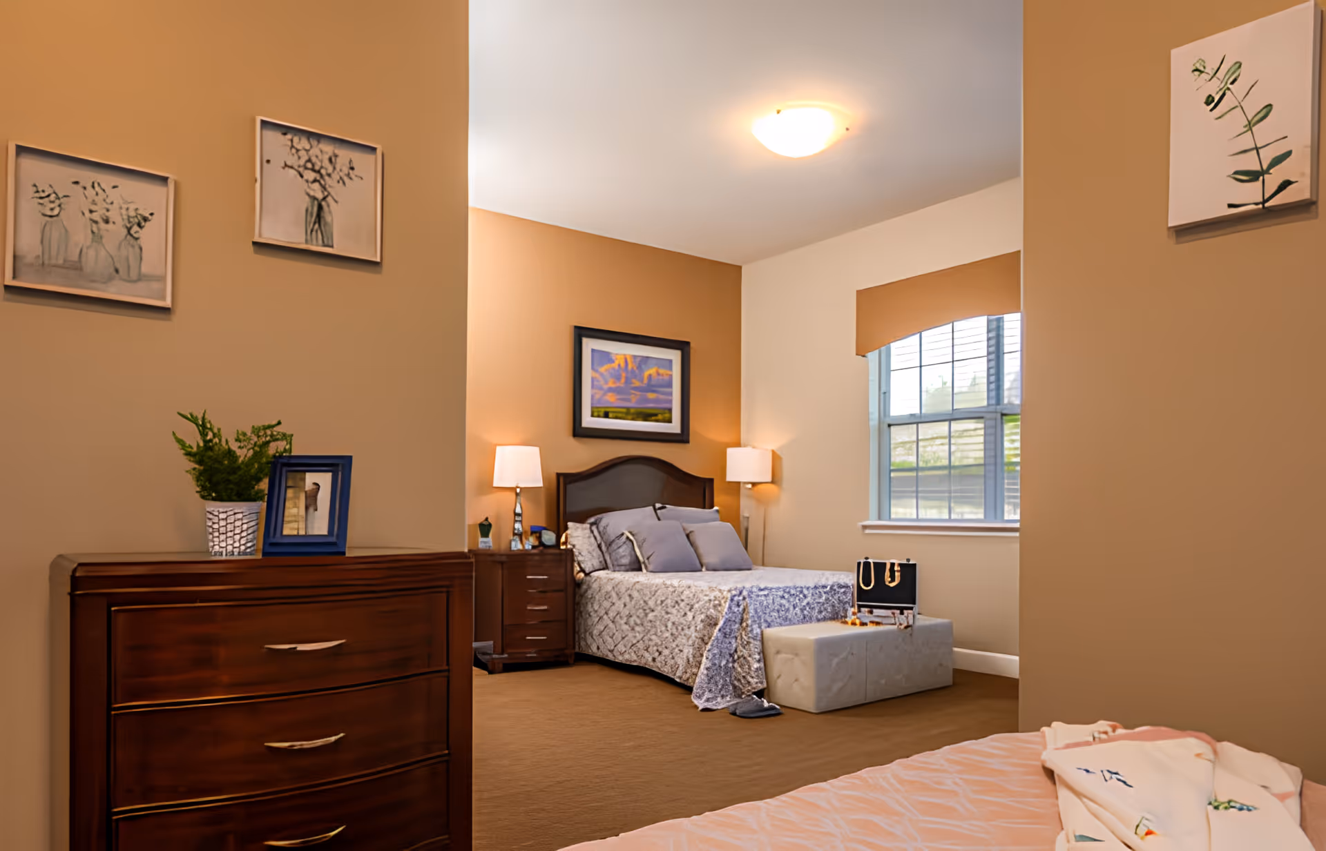 A cozy bedroom in a memory care community with a bed featuring a patterned bedspread and several pillows. There are two bedside tables with lamps on either side of the bed, a window with a valance, a cushioned bench at the foot of the bed with a jewelry box on top, and a wooden dresser with a plant and framed photo. The walls are painted in warm beige tones and decorated with framed artwork.