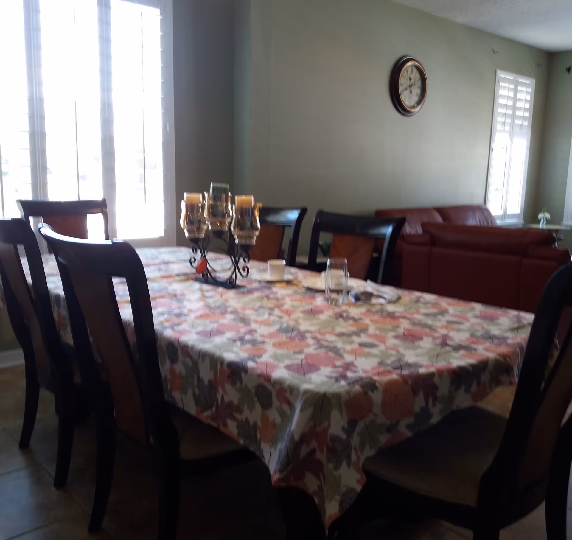 A dining room with a rectangular table covered by a floral tablecloth. Six wooden chairs surround the table. On the table, there is a decorative candle holder with multiple candles, a glass, and a small cup. In the background, there is a red sofa, a wall clock, and windows with white shutters allowing natural light into the room.