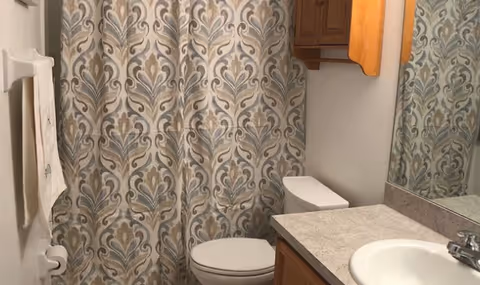 A bathroom with a patterned shower curtain in shades of blue, gray, and beige. There is a white toilet next to a countertop with a sink and a faucet. Above the sink is a large mirror, and a wooden cabinet is mounted on the wall. A white towel hangs on a towel rack on the left side.