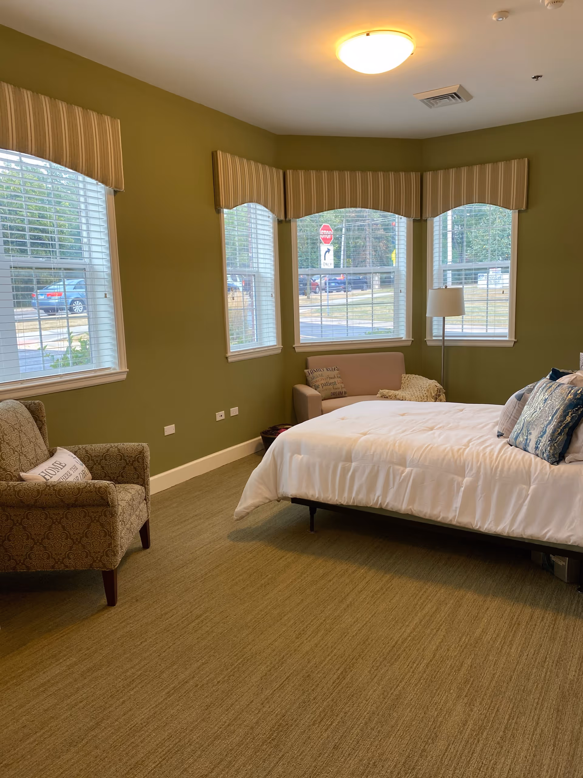 A cozy bedroom with green walls and carpet, featuring a large bed with white bedding and decorative pillows. There is a patterned armchair with a pillow, a small sofa with a throw blanket, a floor lamp, and three large windows with striped valances letting in natural light. Outside the windows, a street with cars and a stop sign is visible.