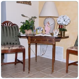 A small wooden table with decorative items including a lamp, a pink angel figurine, a potted plant, a photo frame, and a vase with white flowers. The table is positioned against a wall with a vent below it. Two wooden chairs with cushioned seats flank the table on either side.