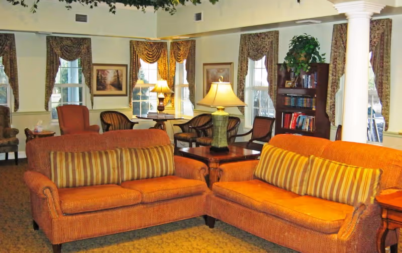 A cozy common room with two orange sofas with striped pillows, side tables and lamps, chairs, a bookcase, and curtained windows.