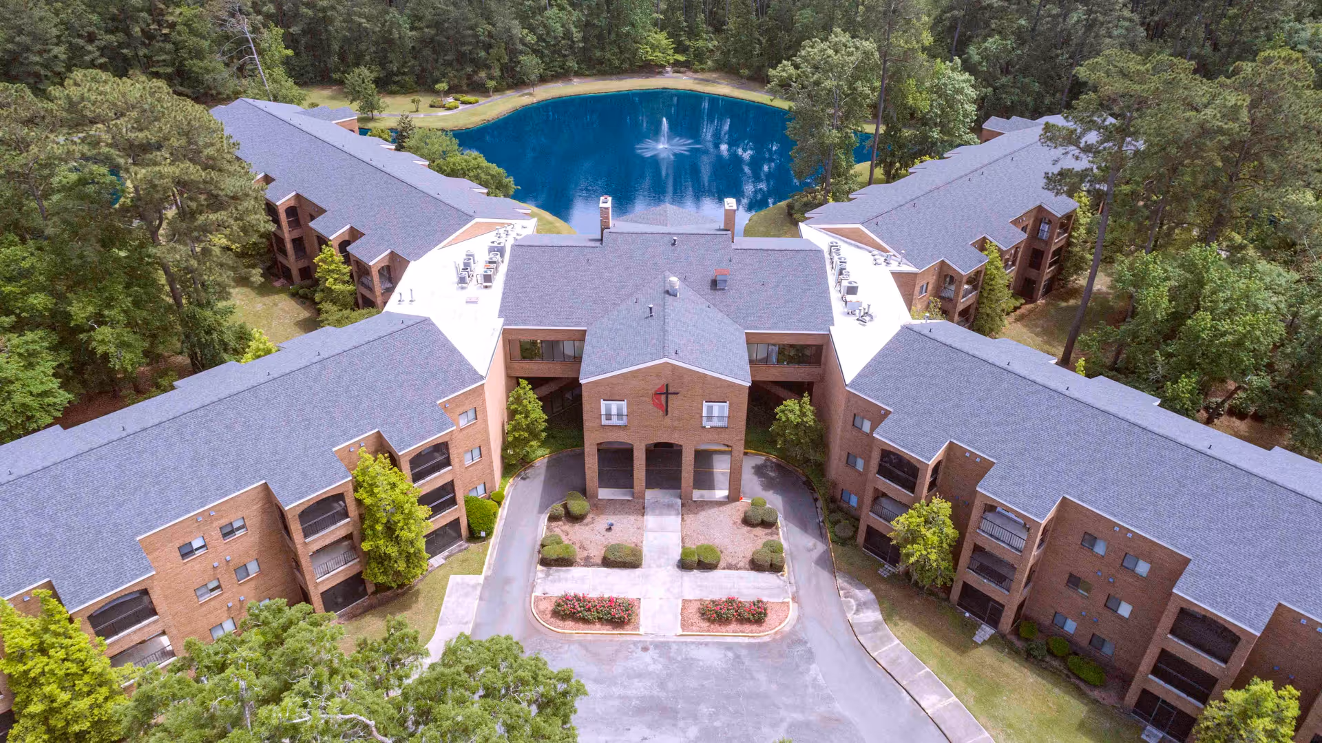 Aerial view of Magnolia Manor of Richmond Hill, showing a large brick building complex surrounding a circular driveway and a central pond with a fountain, all set within a wooded area.