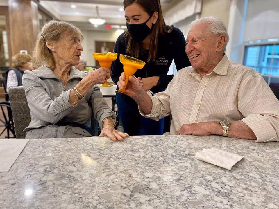 Two elderly individuals sitting at a marble table clinking orange drinks in margarita glasses, with a smiling staff member wearing a black mask and a 'The Preserve' jacket standing behind them in a well-lit dining area.
