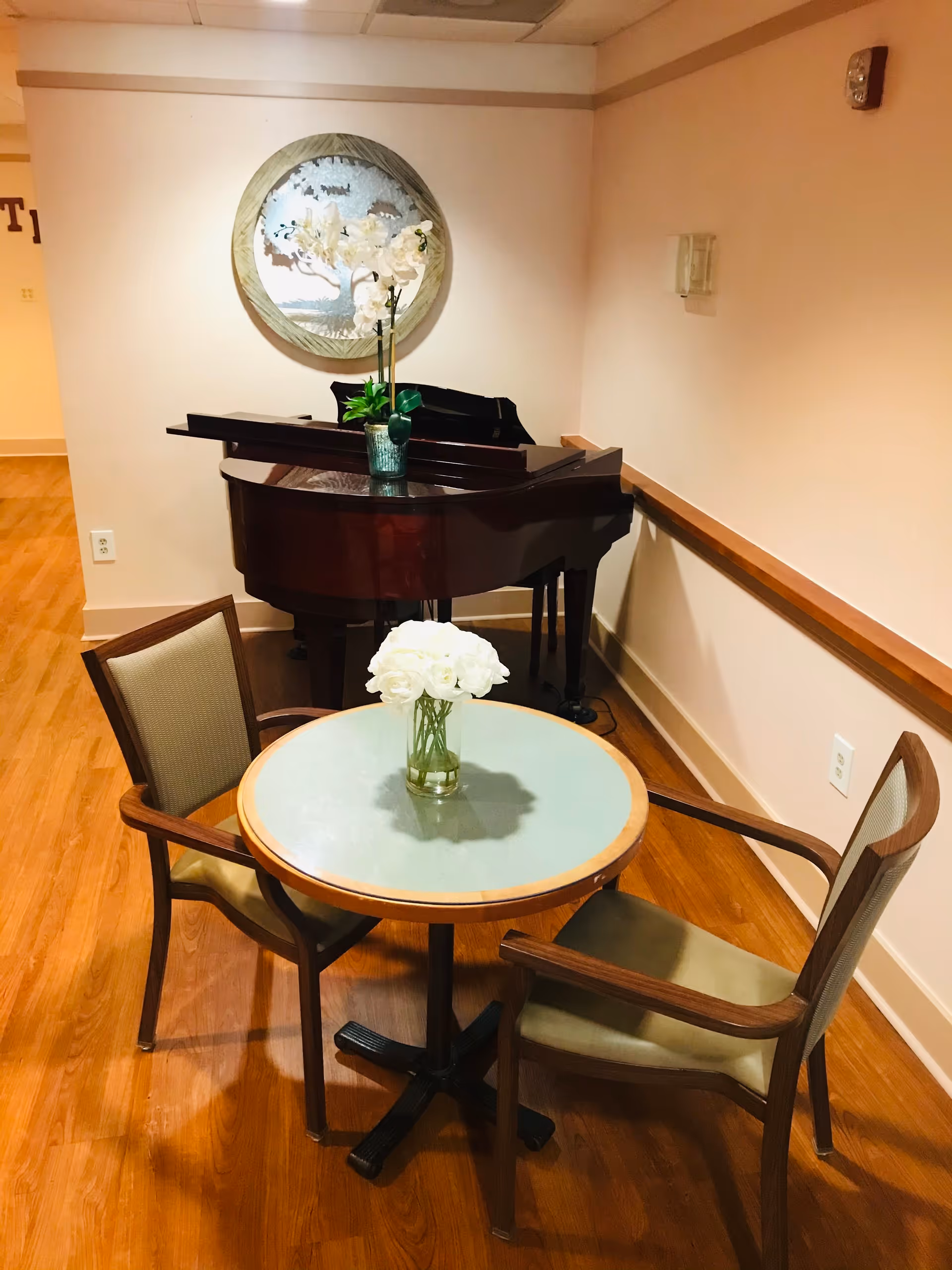 A small round table with a glass top and two wooden chairs with cushioned seats are placed on a wooden floor. A vase with white flowers is on the table. Behind the table is a dark brown grand piano with a small green plant on top. A round decorative wall piece hangs on the light-colored wall above the piano.