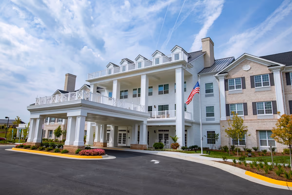 Exterior view of a large, elegant senior living facility building with white columns, multiple windows, and a covered entrance. The building has a mix of white siding and light brick, with an American flag displayed near the entrance. The sky is partly cloudy.