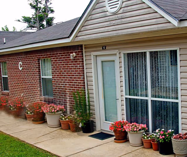 Exterior view of a residential building with a brick and siding facade. There are several potted plants with flowers lined up along the concrete walkway in front of the building. A white door with a window and a large window with curtains are visible. The building has a sloped roof and a small vent near the roof peak.