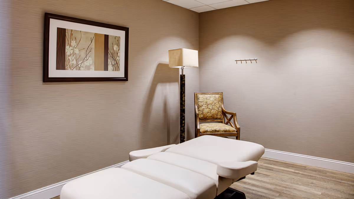 A small treatment room with a padded massage table, upholstered chair, floor lamp, and framed artwork on neutral walls.