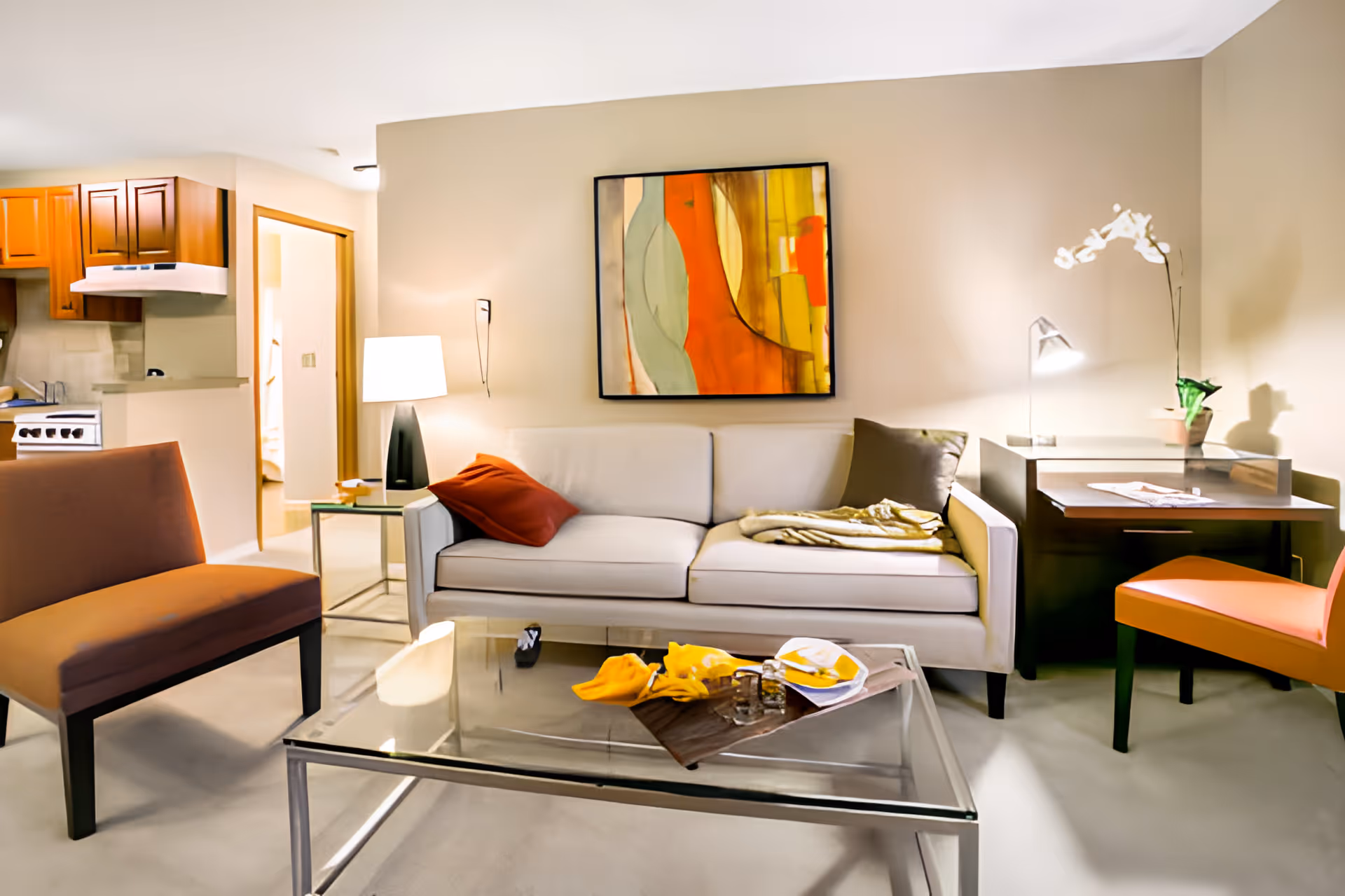 A cozy living room area with a white sofa adorned with a red and a gray pillow, a glass coffee table with decorative items, an orange chair, a small desk with a lamp and a white orchid plant, and a modern abstract painting on the beige wall. The kitchen area with wooden cabinets and a stove is visible in the background.