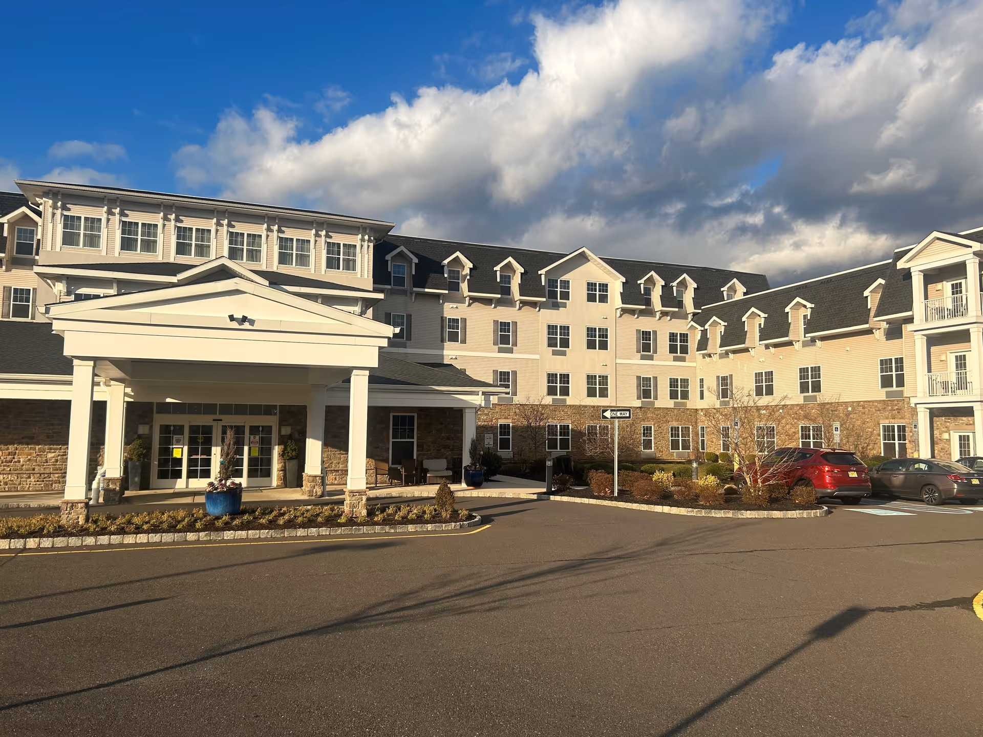 Exterior view of Volante Senior Living of Hamilton, showing a large multi-story building with beige siding and stone accents. The entrance has a covered portico with white columns. Several cars are parked in front of the building under a partly cloudy sky.