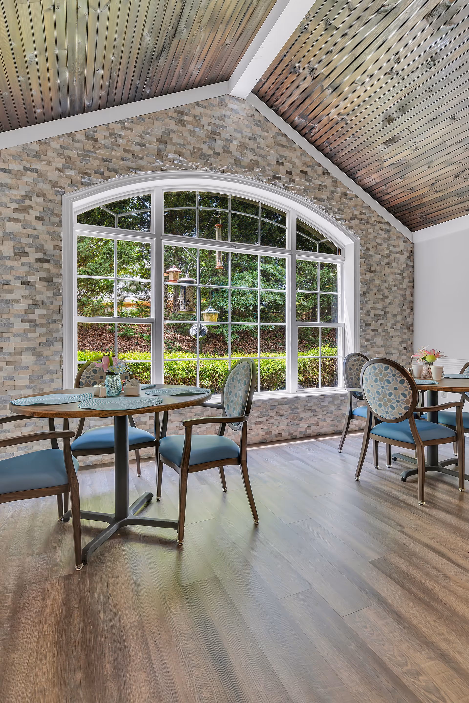 A bright dining area with wooden floors and a vaulted wooden ceiling. The room features a large arched window with multiple panes, looking out onto a green garden with bird feeders. There are round tables with blue cushioned chairs arranged around them, each table set with placemats and small flower vases.