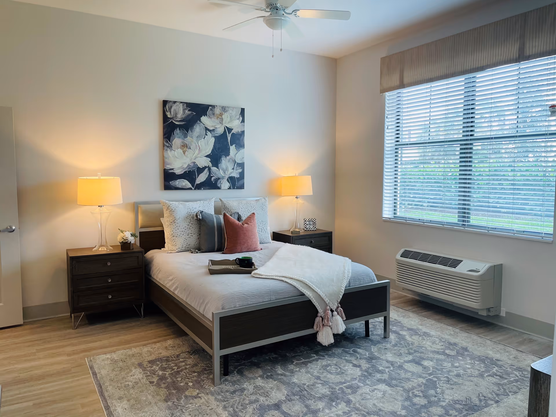A neatly arranged bedroom with a bed centered against a wall, flanked by two dark wooden nightstands each with a lamp. The bed has white and gray bedding with decorative pillows and a white throw blanket. Above the bed is a floral painting. A large window with blinds is on the right wall, and a ceiling fan is visible above. The floor has a patterned area rug.