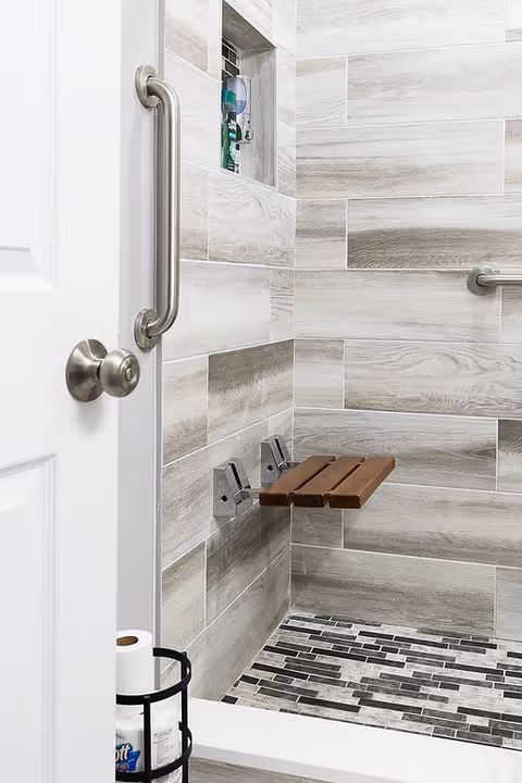 A modern shower area with light gray and beige tiled walls and a mosaic tiled floor. There is a foldable wooden shower seat mounted on the wall, a stainless steel grab bar, and a built-in niche holding shower products. A white door with a round metal knob is partially open, and a black metal holder with toilet paper rolls is visible near the door.