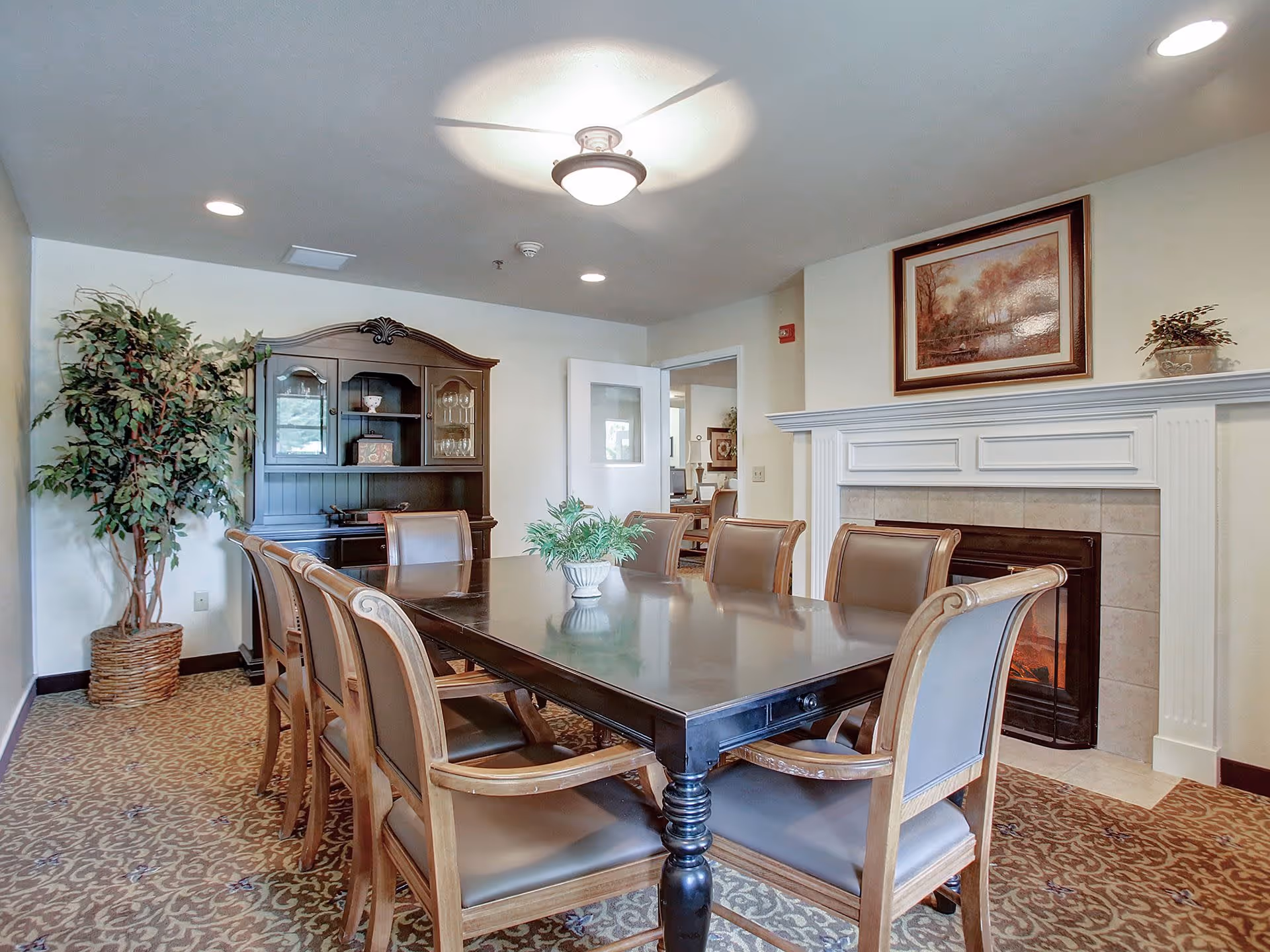 A dining room with a long dark wooden table surrounded by eight cushioned chairs. There is a fireplace with a framed painting above it on the right wall. A large wooden cabinet with glass doors is against the back wall, and a potted plant is in the corner. The room has patterned carpet and recessed lighting in the ceiling.