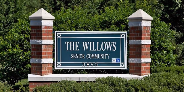 A green and white sign reading 'THE WILLOWS SENIOR COMMUNITY 3850' mounted between two brick pillars with greenery and bushes surrounding it.