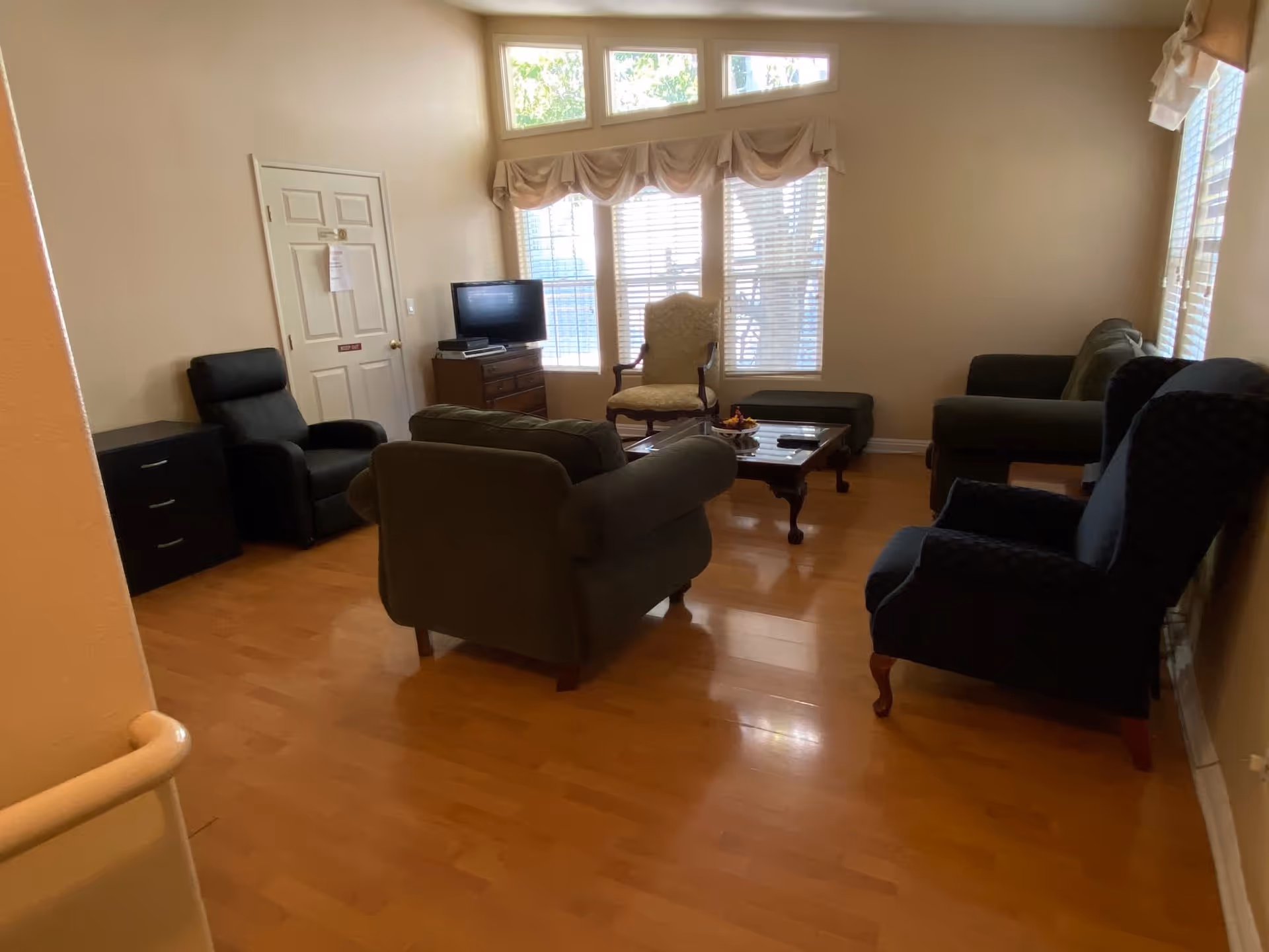 A cozy living room with wooden flooring, featuring multiple armchairs and sofas arranged around a wooden coffee table. A small TV sits on a wooden dresser near a window with blinds and decorative valances. The room is softly lit by natural light coming through the windows.