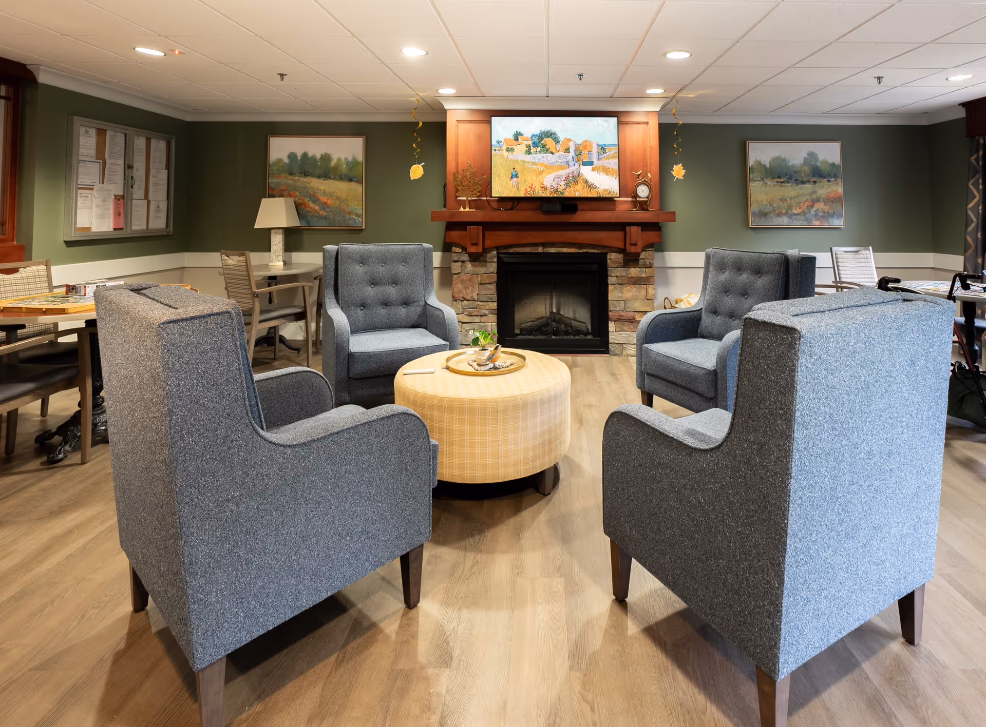 A cozy common area with four gray upholstered armchairs arranged around a round, yellow plaid ottoman. Behind the seating area is a stone fireplace with a wooden mantle, above which hangs a colorful painting. The walls are painted green with white trim, and there are additional tables and chairs along the walls. Two landscape paintings and a bulletin board are visible on the walls.
