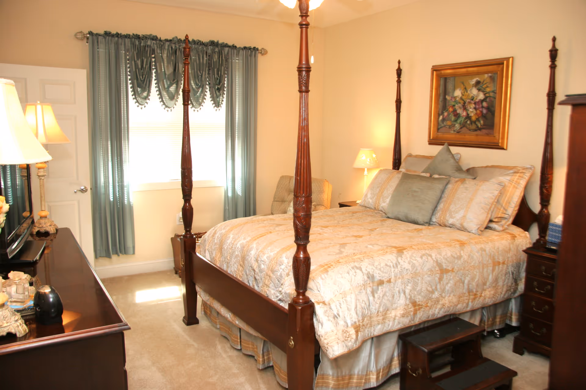 A cozy bedroom with a wooden four-poster bed dressed in beige and gold bedding, several pillows, and a small wooden step stool at the foot of the bed. The room has beige walls, a window with teal curtains, a wooden dresser with a lamp and decorative items, a framed floral painting on the wall, and a cushioned chair in the corner.