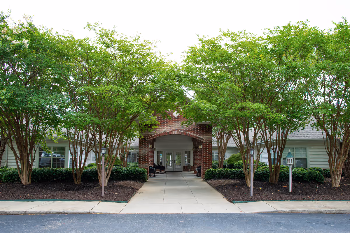 Entrance to a senior living facility with a brick archway, a concrete walkway, and green trees and shrubs on either side.