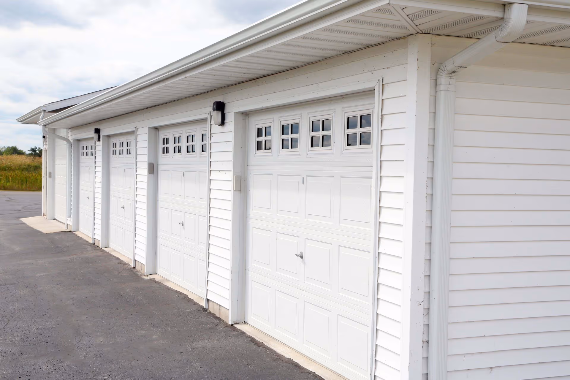 Exterior view of a white building with four closed white garage doors, each having small square windows at the top. The building has white siding and a paved driveway in front.
