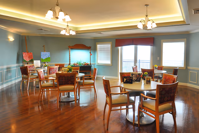 A bright and spacious dining room with several round tables and wooden chairs with patterned cushions. The room has light blue walls, large windows with blinds and a red valance, and polished wooden floors. There are ceiling lights and wall sconces providing warm lighting. Some tables have small flower arrangements and framed photos, and there are two easels with paintings near the back wall.