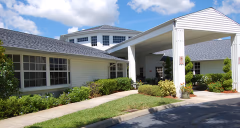 Exterior view of a single-story building with white siding and a covered entrance. The building is surrounded by green shrubs and a well-maintained lawn under a partly cloudy blue sky.