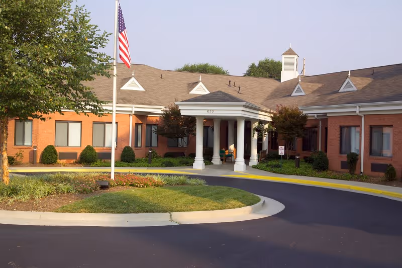 Exterior view of a single-story brick building with a covered entrance supported by white columns. There is a circular driveway with a landscaped island in the center, featuring grass, bushes, and a tree. An American flag is flying on a flagpole near the entrance.