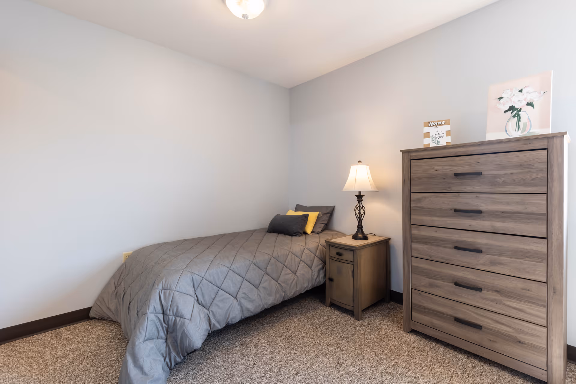 A small bedroom with a single bed covered in a gray quilt, two pillows, a wooden nightstand with a decorative lamp, and a wooden chest of drawers with a small framed sign and a painting of flowers on top. The walls are light gray and the floor is carpeted.