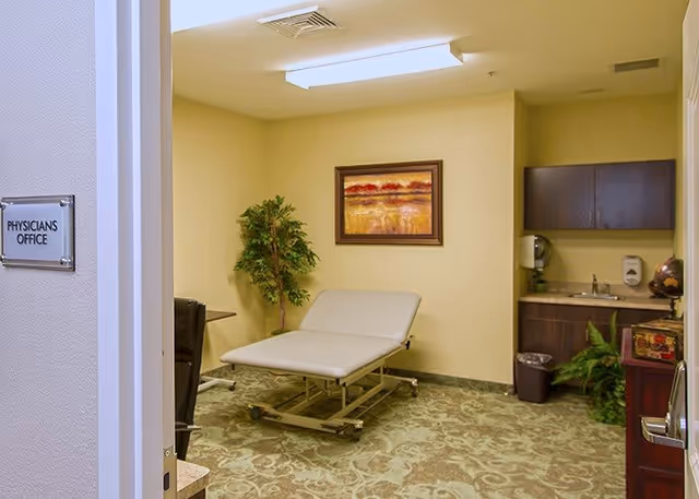 Interior view of a physician's office with a medical examination table, a framed landscape painting on the wall, a potted plant, a small sink area with cabinets, and a trash bin. The room has beige walls and patterned carpet flooring.
