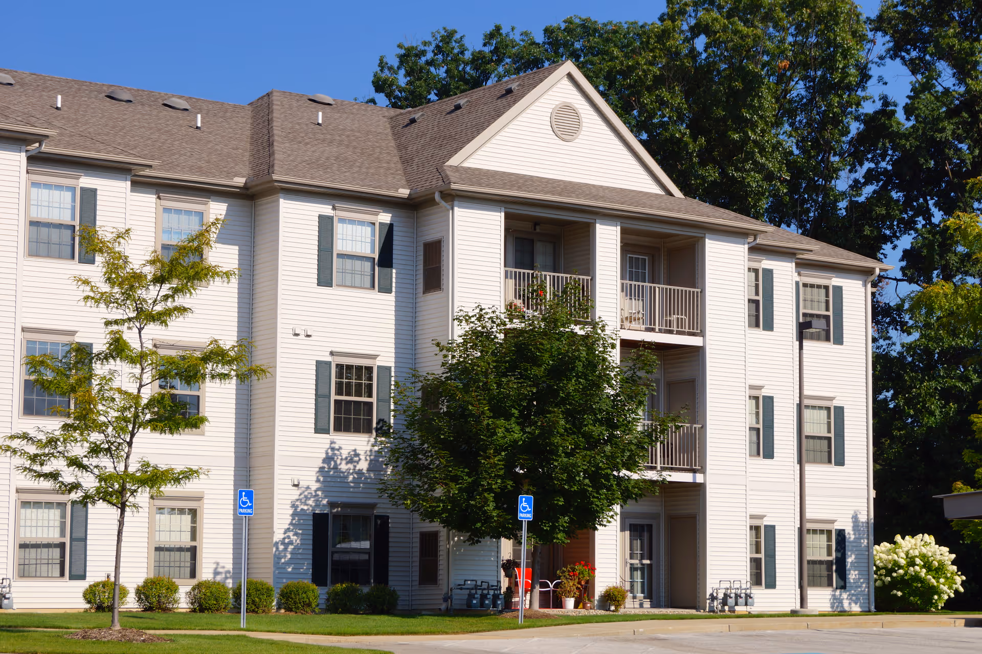 Three-story white residential building with balconies, trees, and a parking area with handicap signs.