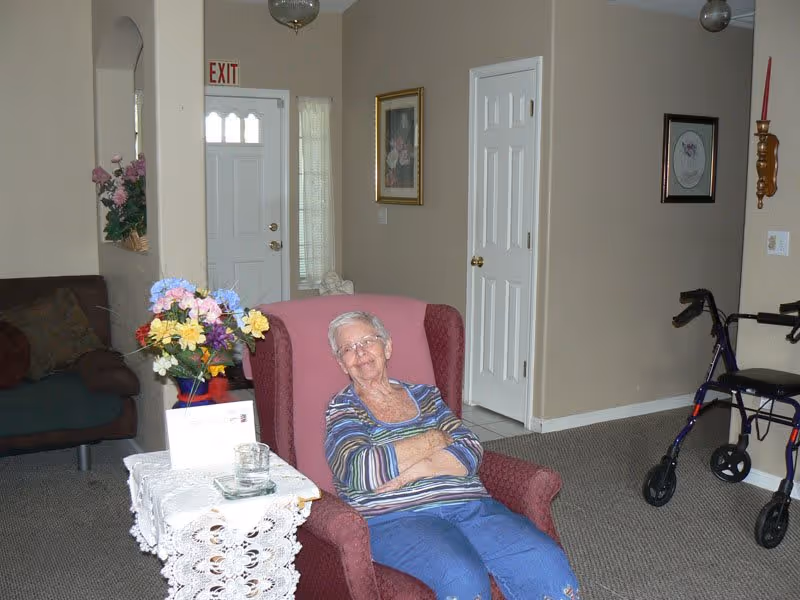 An elderly woman with short white hair and glasses sits comfortably in a red armchair in a living room. She is wearing a striped long-sleeve shirt and blue pants. Next to her is a small table covered with a white lace cloth, holding a colorful bouquet of flowers and a glass ashtray. In the background, there is a brown couch, a white door with an exit sign above it, framed pictures on beige walls, and a blue walker with black handles on the carpeted floor.