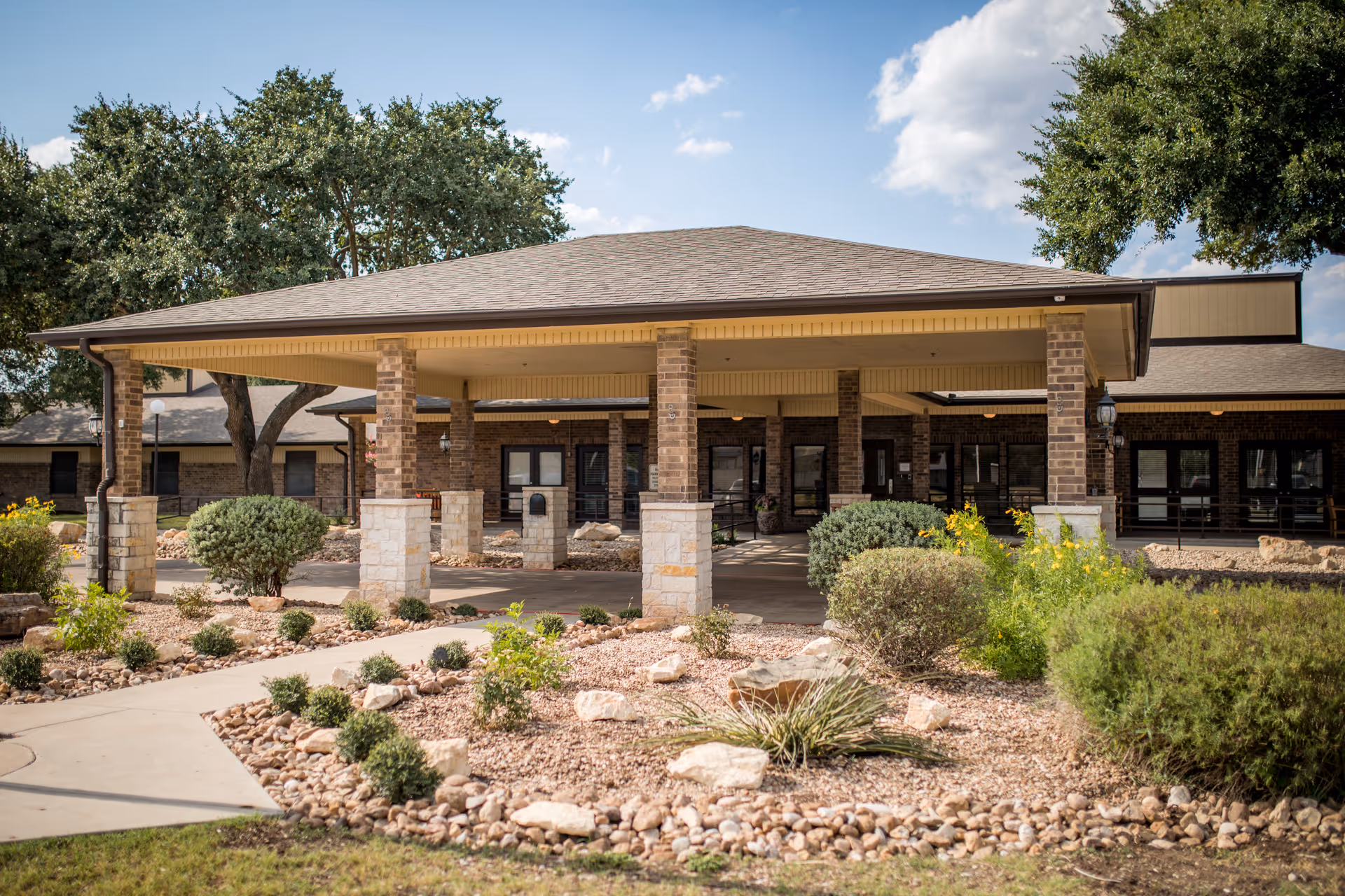 Exterior view of Silver Pines Nursing and Rehabilitation Center showing the entrance with a covered drop-off area supported by brick columns, surrounded by landscaped garden beds with rocks, shrubs, and small plants under a partly cloudy sky.