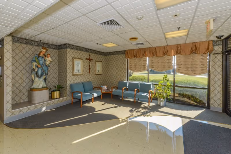 A waiting area with blue cushioned chairs arranged around a small wooden table. The walls have patterned wallpaper and religious decorations including a statue of the Virgin Mary holding baby Jesus, a crucifix, and framed pictures. Large windows with beige curtains allow natural light to fill the room, and there are a couple of potted plants near the windows.