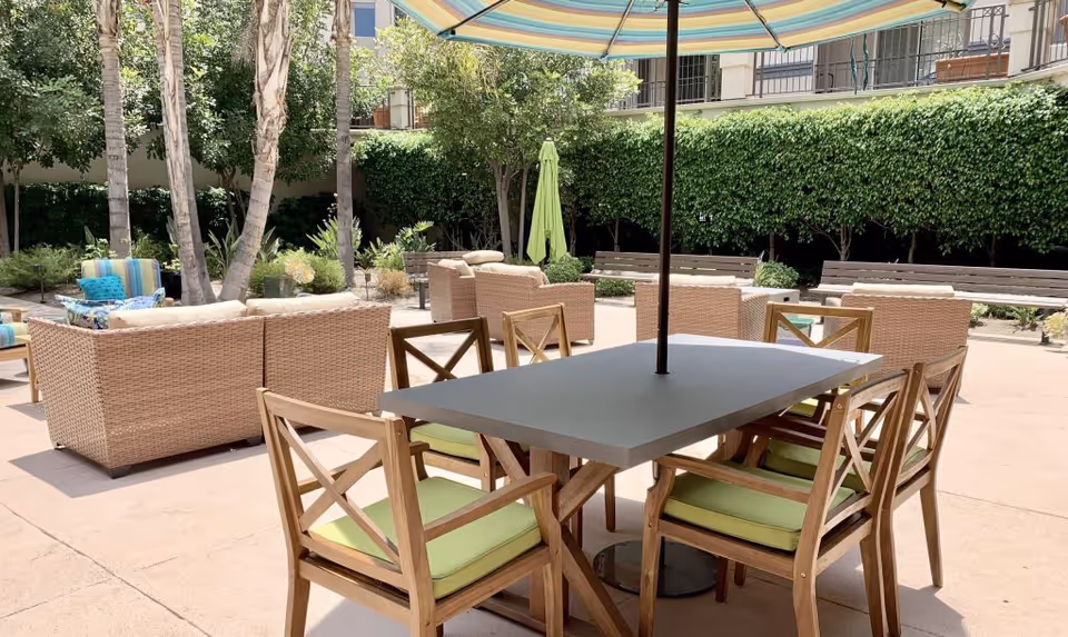 Outdoor patio area with a rectangular table surrounded by six wooden chairs with green cushions. A large striped umbrella shades the table. In the background, there are wicker sofas with cushions, trees, and a tall hedge providing privacy.