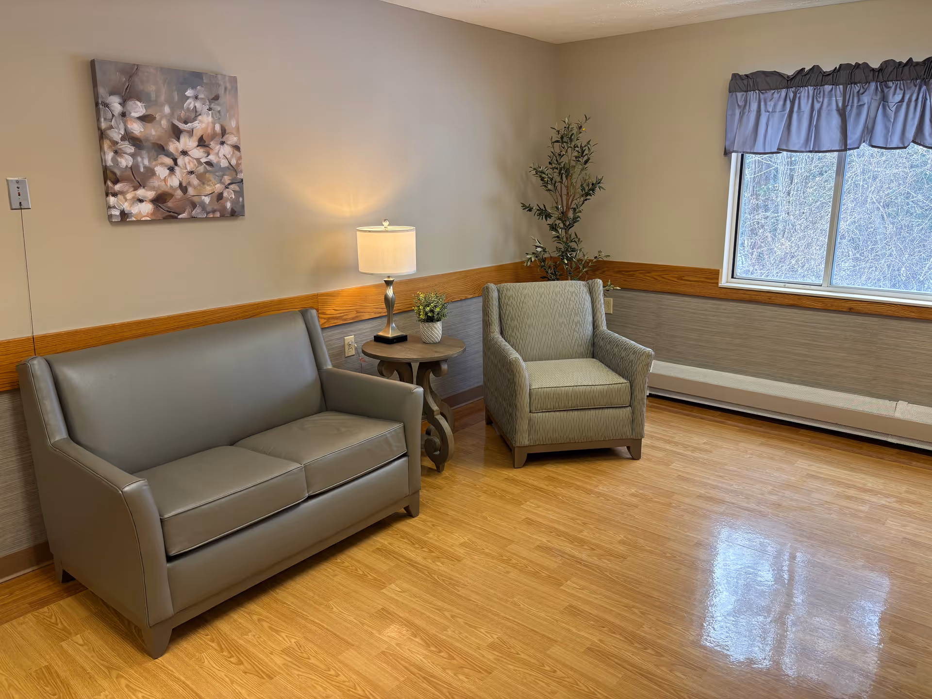 A small sitting area in a room with a gray leather loveseat, a patterned armchair, a round wooden side table with a lamp and a small plant, a large window with a blue valance, a potted artificial tree in the corner, and a floral painting on the wall. The floor is wooden and the walls are beige with a wooden chair rail.