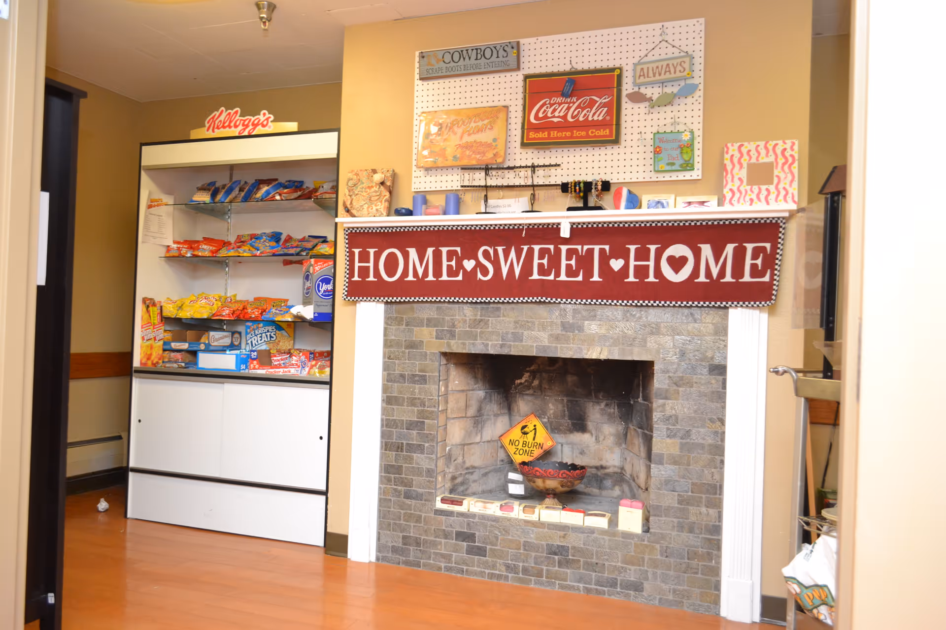 Interior view of a cozy room with a stone fireplace decorated with a maroon banner that reads 'HOME SWEET HOME'. Above the fireplace are various vintage-style signs and decorations. To the left of the fireplace is a white shelving unit stocked with assorted snacks and Kellogg's branding on top. The room has light brown walls and wooden flooring.