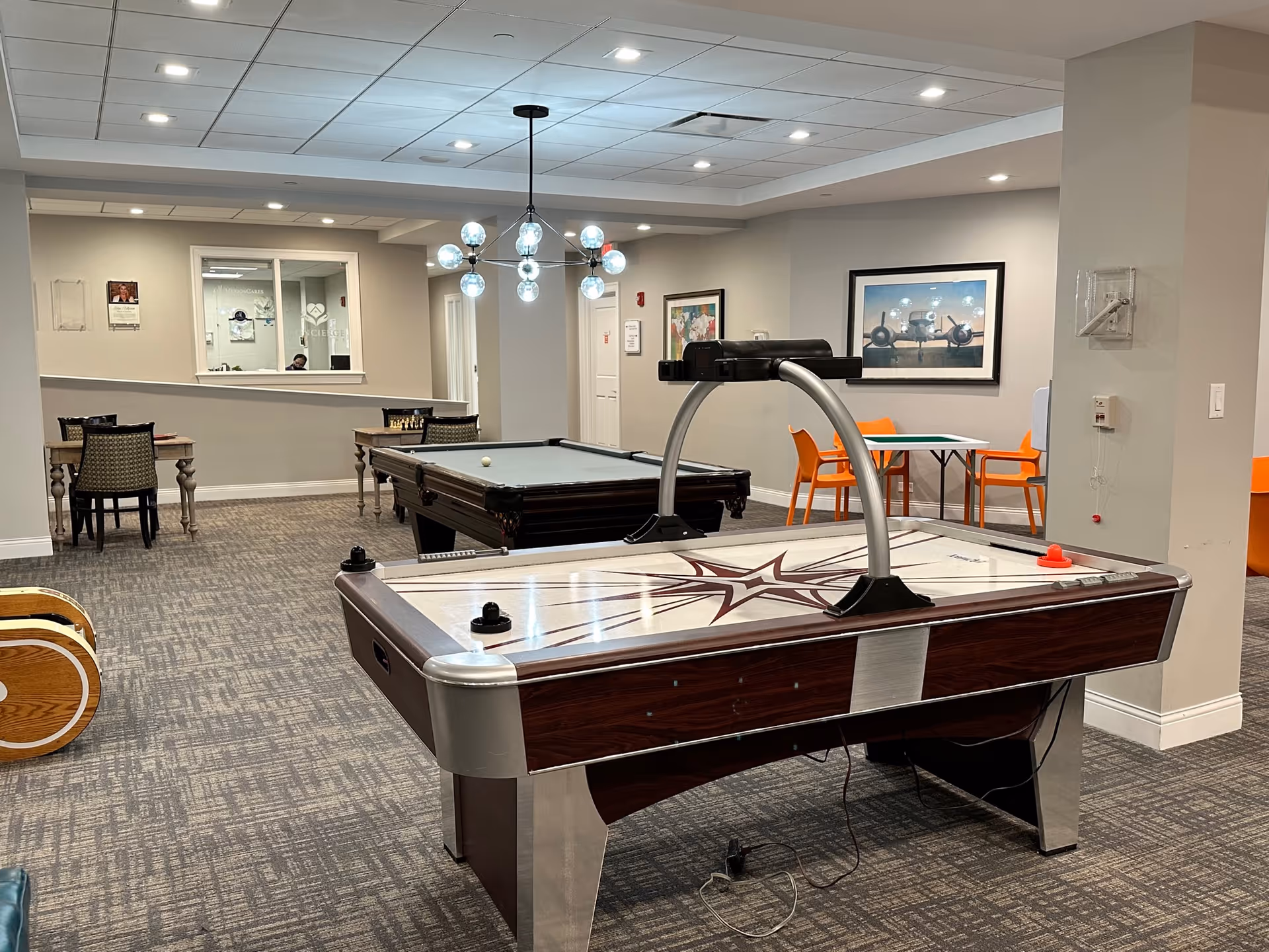 A recreational room with an air hockey table in the foreground and a pool table behind it. There are several tables and chairs around the room, including a pair of orange chairs at a small table near a wall with framed artwork. The room has a carpeted floor, neutral-colored walls, and a modern light fixture hanging from the ceiling.