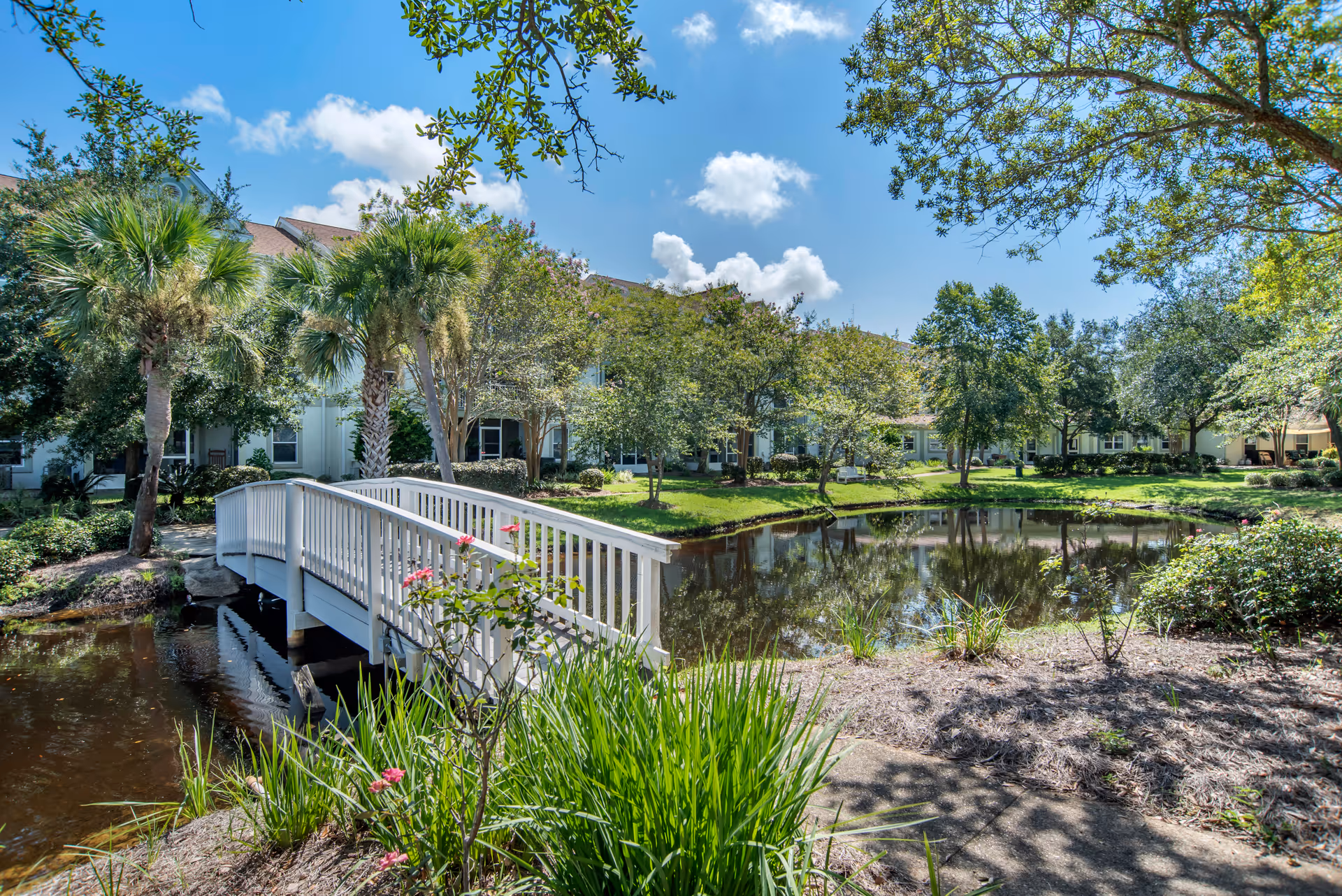 A white footbridge crosses a pond in a landscaped courtyard with palm trees and residential buildings under a blue sky.
