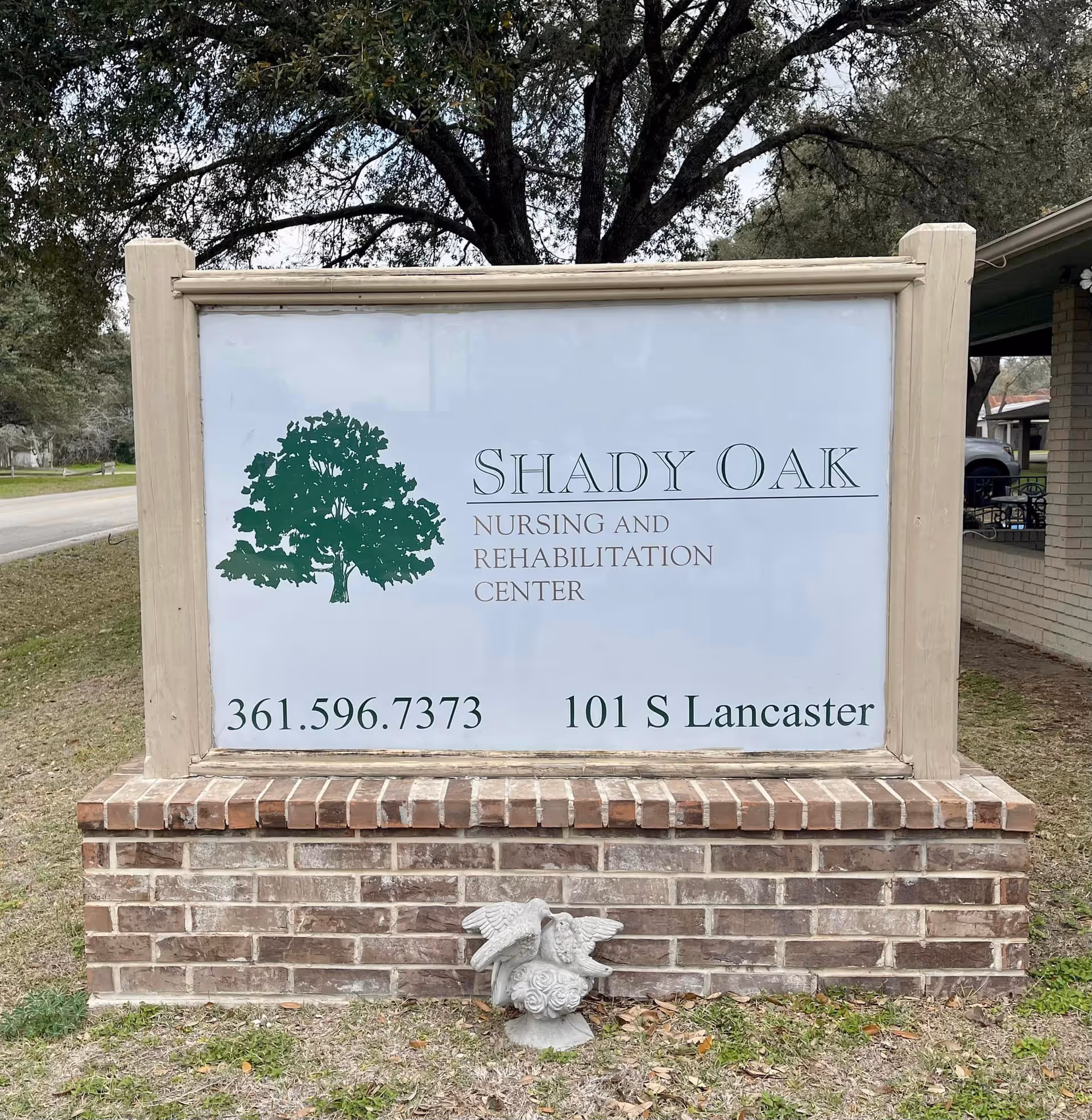 Outdoor sign for Shady Oak Nursing and Rehabilitation Center with a green tree logo, phone number 361.596.7373, and address 101 S Lancaster, set on a brick base with a small decorative statue of two birds in front.