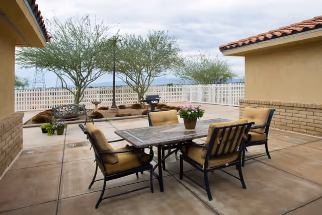 Outdoor patio area with a rectangular table and four cushioned chairs. A potted plant with pink flowers is on the table. The patio is surrounded by a white picket fence, with trees and a bench in the background under a cloudy sky.