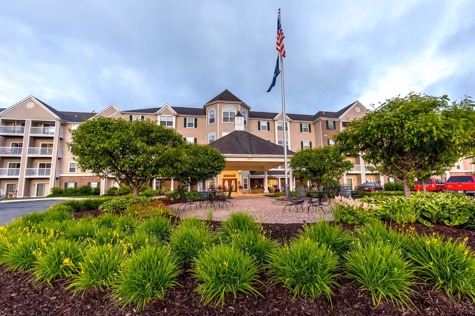 Exterior view of Independence Village Of Petoskey, showing a multi-story building with balconies, a covered entrance, and a flagpole with American and state flags. The foreground features landscaped gardens with green shrubs, small trees, and outdoor seating on a paved area.