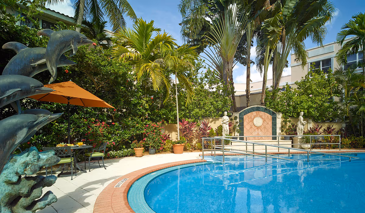 Outdoor swimming pool area with a dolphin statue fountain on the left, surrounded by tropical plants and palm trees. There is a table with chairs and an orange umbrella near the pool, and a decorative wall with statues in the background under a partly cloudy sky.