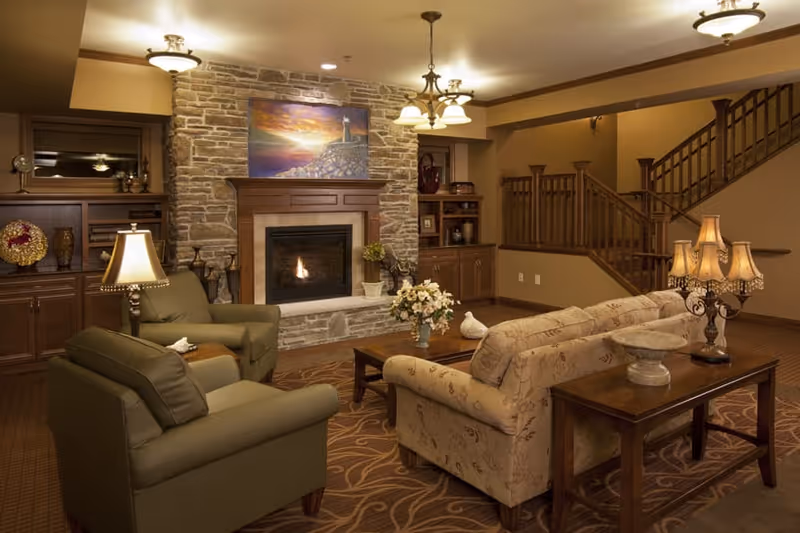 A cozy living room with a stone fireplace in the center, a painting above it, two armchairs and a floral-patterned sofa arranged around a wooden coffee table with a flower vase. There are built-in wooden shelves and cabinets on either side of the fireplace, a staircase with wooden railing in the background, and warm lighting from ceiling fixtures and table lamps.
