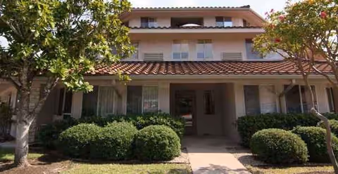 Two-story stucco building with a tiled roof, shrubs, and a walkway to the entrance.